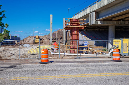Highway bridge underpass undergoing construction in St. Petersburg, FL with orange traffic cones, barriers, work vehicles and equipment on a sunny day, showing road repair, safety measures and urban i