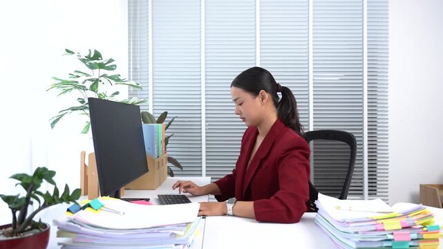 Focused Professional: A diligent woman works intently at her computer in a bright, modern office, surrounded by stacks of paperwork and potted plants.