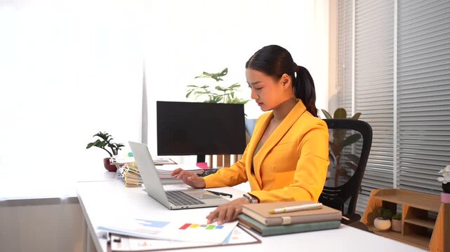 Focused Businesswoman at Desk: A concentrated businesswoman in a vibrant yellow blazer works diligently at her modern office desk, analyzing reports on her laptop with a stack of books beside her.