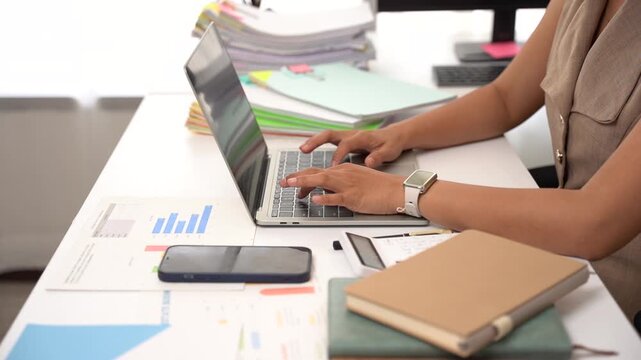 Focused Workstation: A woman diligently works on her laptop in a bright office, surrounded by stacks of documents, charts, and office supplies, signifying a busy workday.