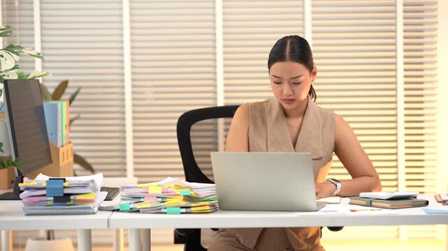 Focused Professional at Work: A diligent woman is engrossed in her work at a desk, surrounded by stacks of documents and a laptop, under the soft glow of office lighting.