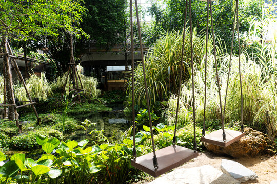Two empty wooden swings hanging by ropes in a lush green tropical garden with a small pond and cafe