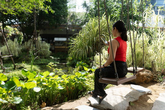 An Asian woman in a red top sitting on a wooden rope swing and using her smartphone in a lush green tropical garden with a pond