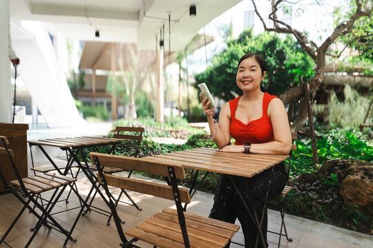 Portrait of a smiling Asian woman in a red top sitting at an outdoor wooden cafe table holding a smartphone with a lush green garden background