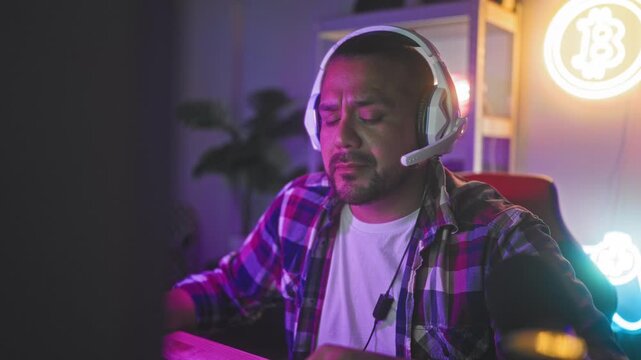 Man in headset and plaid shirt pinches nose and closes eyes at computer desk in studio during gaming session; frustration fatigue.