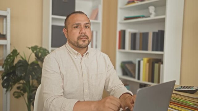 Man at laptop with stack of folders and bookshelf makes finger to lips for silence in a building office; secrecy focus.