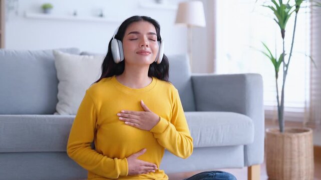 Asian woman meditates and practices Zen meditation at home, focusing on breathing exercises and seeking spiritual peace in her living room. She sits on the floor, listening podcast to headphones