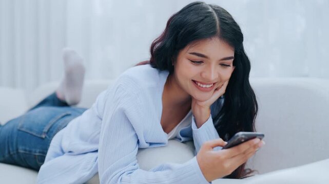 Young Asian woman using her cellphone while relaxing on the sofa at home, typing chat message on smartphone, smiling girl use cell phone chatting, searching information on browser wireless internet