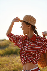 Fototapeta premium Young woman enjoying summer lifestyle outdoors. She wears a striped shirt and straw hat, holding a basket. Sunset light, concept for vacation, freedom, leisure and joy.