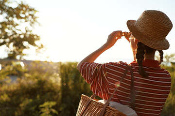 Fototapeta premium Young woman in hat taking pictures with camera outdoor. Lifestyle portrait with summer sun flare. Concept of travel, photography, hobby, and relaxation in nature.