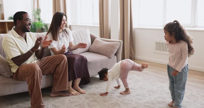 Little biracial sisters toddler and preschooler performing funny song and dance for proud multiethnic parents in living room getting support encouragement applause as reward for joyful domestic show
