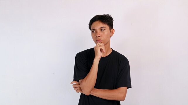 Young Asian man touching his chin with a doubtful or hesitant look, glancing sideways against a white background.