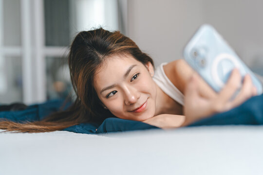 Happy young adult asian woman waking up in bedroom on morning enjoy checking smartphone social media Relax rest and leisure lifestyle