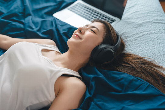 Mindfulness young adult asian woman lying in bed, wearing headphones listening relax music in cozy comfort bedroom