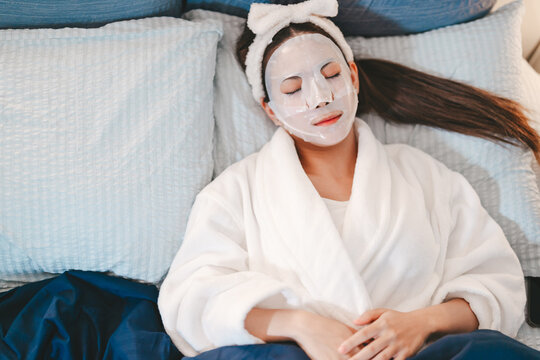 Relax young adult asian female wearing bathrobe with beauty face mask closed eyes, sleeping on bed