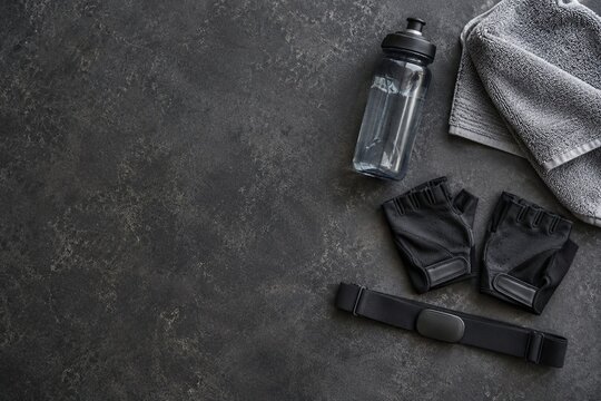 Water bottle, fingerless gloves, gray towel with heart rate chest strap on dark stone surface, cycling workout gear flat lay