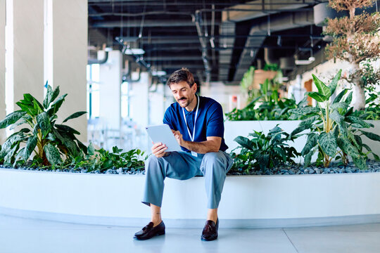 Man with lanyard using a digital tablet, sitting on a planter in a green modern office space, remote working