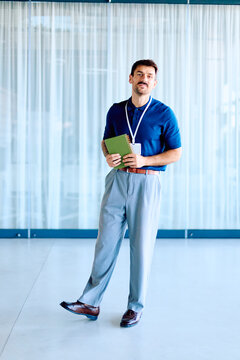 Man in business casual attire holding a notebook and ID badge, standing indoors with a confident expression