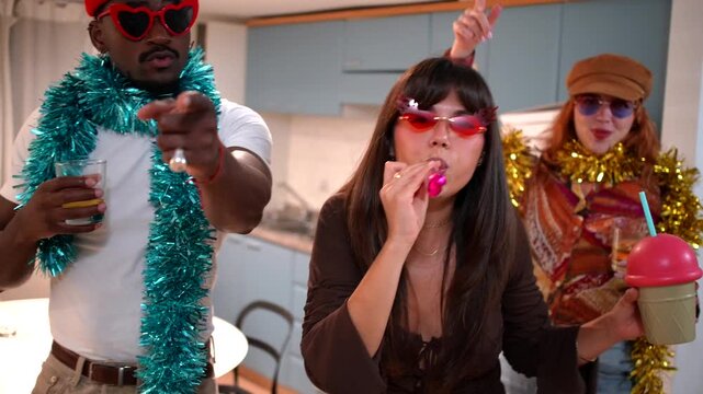 Cheerful multiethnic friends wearing funny glasses and tinsel boas dancing and enjoying drinks together during a vibrant and festive celebration in an apartment kitchen