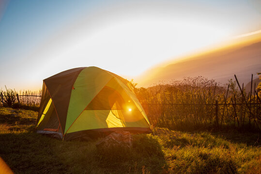 Tent camp in nature background with sunrise on top of mountain. Tent Camping trip outdoor hiking mountain hill. Adventure Travel wildness flare sunlight. Picnic tent green nature campground in forest