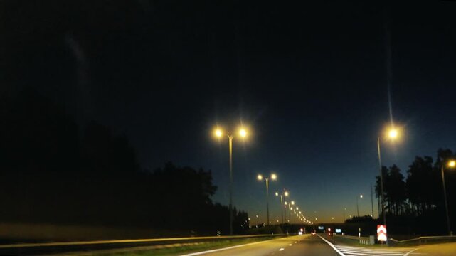 First person perspective from a car traveling on a long, straight motorway at night. Street lamps illuminate the dark road ahead, creating a rhythmic pattern of light for the nighttime journey