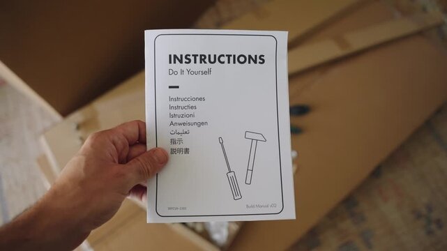 Man's hand holding a Do It Yourself instruction manual over an open cardboard box with flat pack furniture parts, preparing for assembly and home improvement in a new house