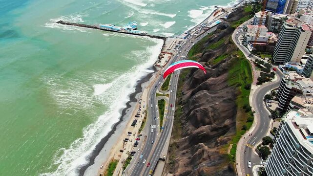 Drone follows a red and white paraglider flying along the Costa Verde cliff edge in Miraflores, Lima, Peru, toward a pier, with beach, waves, highway and seaside towers below.