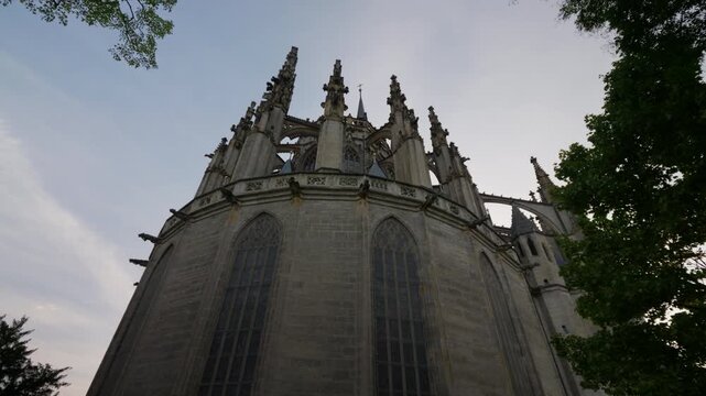 Dynamic side-angle tracking shot of St. Barbara's Cathedral top, showcasing dramatic gothic spires, intricate stone tracery, and sunset sky framed by lush green trees. No people visible.