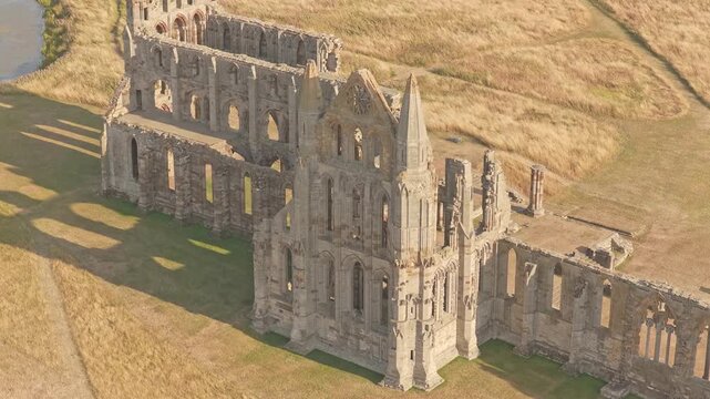 Whitby Abbey medieval Gothic ruins on Whitby Headland, with preserved church walls, pointed arches, transept and surrounding grassland, drone ascent