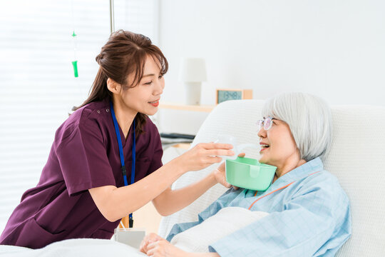介護用コップで水を吸い飲みさせる介護士,看護師の女性と入院患者の高齢者女性