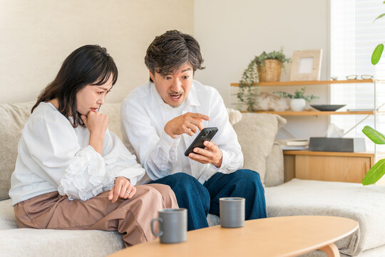 リビングでスマホの画面を見て驚く夫と心配そうな妻。予想外の知らせやトラブルに戸惑う40代・50代夫婦