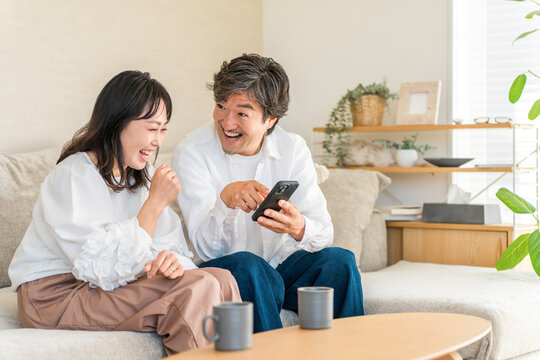 リビングでスマホの面白い動画を見て大笑いする40代・50代の仲良し夫婦。笑顔あふれる楽しい日常