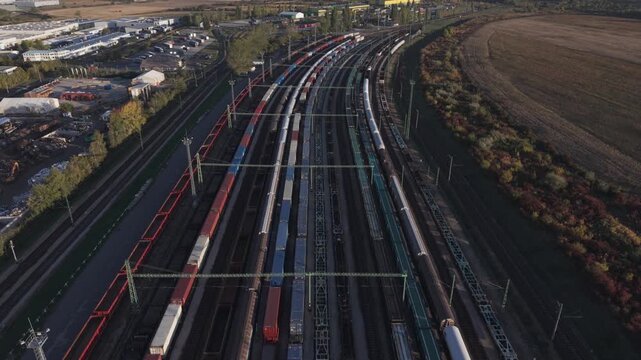 Elevated aerial view of a large railway yard filled with freight trains and cargo containers, bordered by industrial buildings, farmland, and autumn vegetation