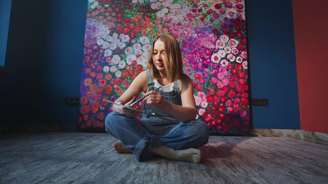 Young artist sits cross-legged, painting with focus. Colors splash behind her, vivid and alive. Palette spins with hues, tools in hand. Peaceful, creative energy fills the room. Ideal for art studios