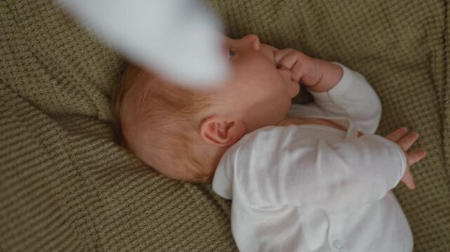 Baby in a bassinet on a knitted cushion with hand to mouth, sucking fingers inside a building; calm comfort.