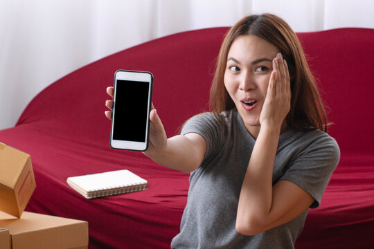 Surprised young Asian female small business owner showing a blank black smartphone screen with an excited expression near parcel boxes and a red sofa at home for ecommerce app mockup.