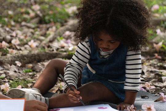 Cute little child with curly hair sitting on a mat in an outdoor park, holding a pencil to draw and write on paper with toy numbers, enjoying early childhood education and creative learning in nature.