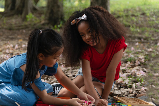 Two happy diverse little girls sitting together in an outdoor park, smiling, drawing, and playing with educational wooden alphabet toys, representing childhood friendship, teamwork, and learning.