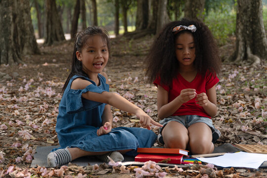 Two diverse little girls sitting on a mat in an outdoor park with books and drawing paper, one happy Asian girl pointing a finger while playing and learning together in childhood friendship.