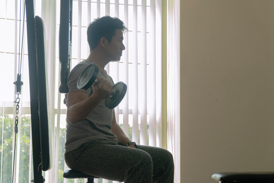 Profile view of a determined young Asian man sitting on a bench in a gym and lifting a dumbbell, focusing on bicep curls, representing strength training, fitness motivation, and an active lifestyle.