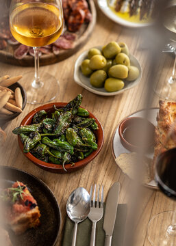 A gourmet spread of Spanish tapas including blistered padron peppers, green olives, cured meats, and picos breadsticks served with a chilled glass of white wine on a wooden table.