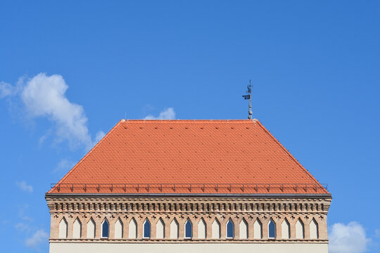 A wide shot of a classic steep orange clay tile roof with a weather vane on top, positioned above a decorative brick cornice of a historic building under a blue sky with soft white clouds in Zagreb