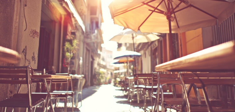 Empty outdoor cafe tables and chairs lining a narrow street. Sunlight creating a warm, inviting atmosphere for relaxation and dining