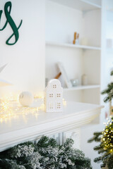 Naklejka premium White festive fireplace mantel with miniature house, glowing lights, green garland, and snowy tree.