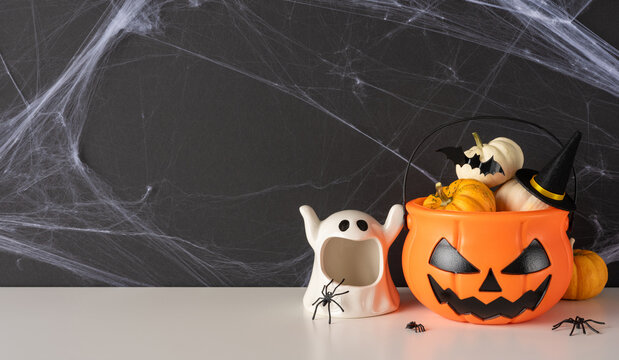 Happy Halloween celebration with pumpkin bucket, ghost decoration, spiders and witch hat on cobweb background on wide banner wall and table background