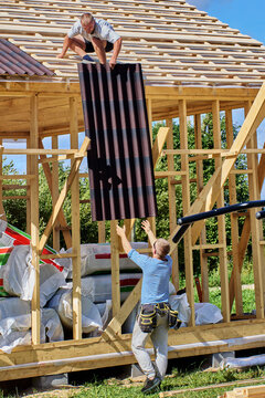 Corrugated bitumen sheeting is passed upward by ground worker to colleague seated on roof slope during installation of roofing on timber frame.