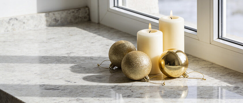 White pillar candles and golden Christmas baubles arranged on a marble surface by a window
