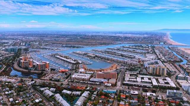 A city marina opens toward the coast between rows of buildings. White boats line parallel piers beneath streaked clouds as a sandy shoreline recedes on the right.