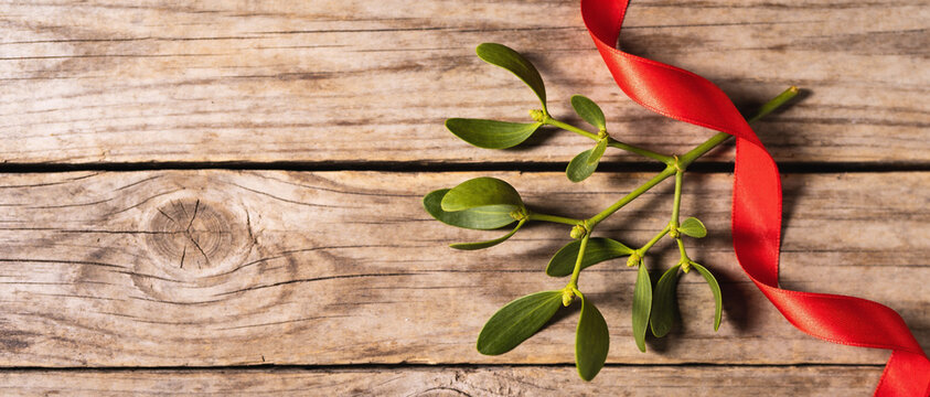 A sprig of mistletoe with green leaves and a red ribbon resting on a rustic wooden surface with white space.