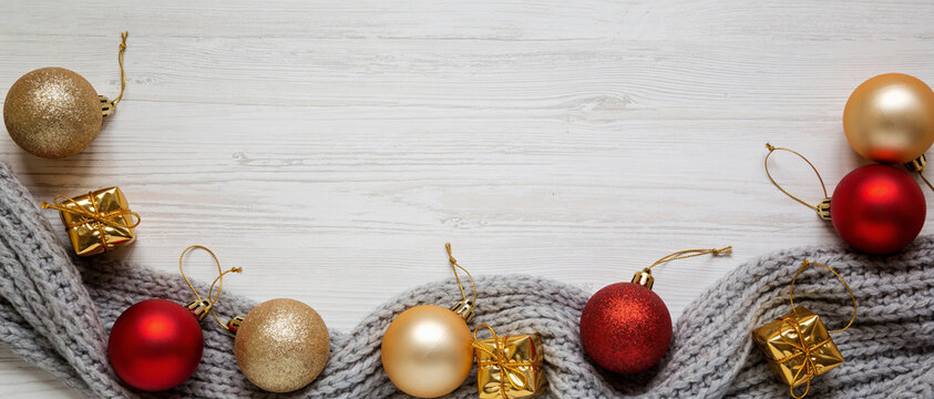 Red, gold, and shiny Christmas baubles and ornaments arranged on a folded gray knitted scarf on a white wooden background.